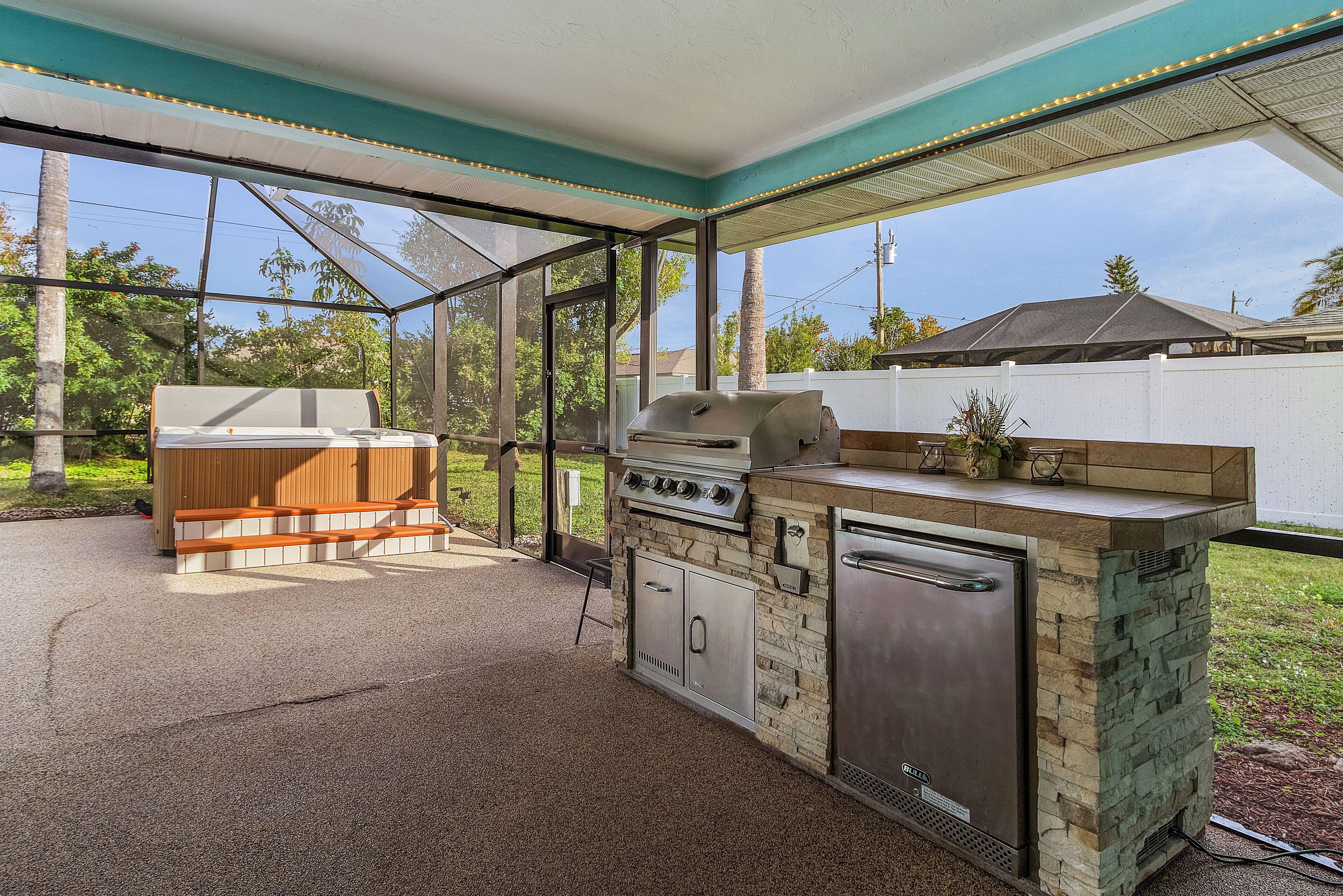 An outdoor kitchen with a grill, oven, and built-in refrigerator, enclosed by a glass structure with a view of the backyard.