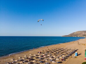 Una playa con tumbonas y sombrillas, cielo despejado y un parapente a lo lejos.