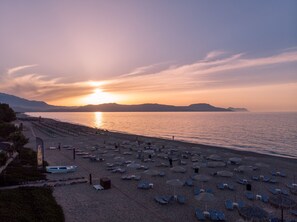 Una playa con filas de tumbonas y sombrillas, un pequeño bote, y una vista despejada del atardecer sobre el agua.