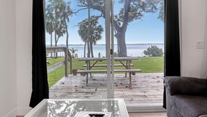 Habitación con un gran ventanal corredera que da a una terraza exterior con mesas de picnic y vistas al agua.
