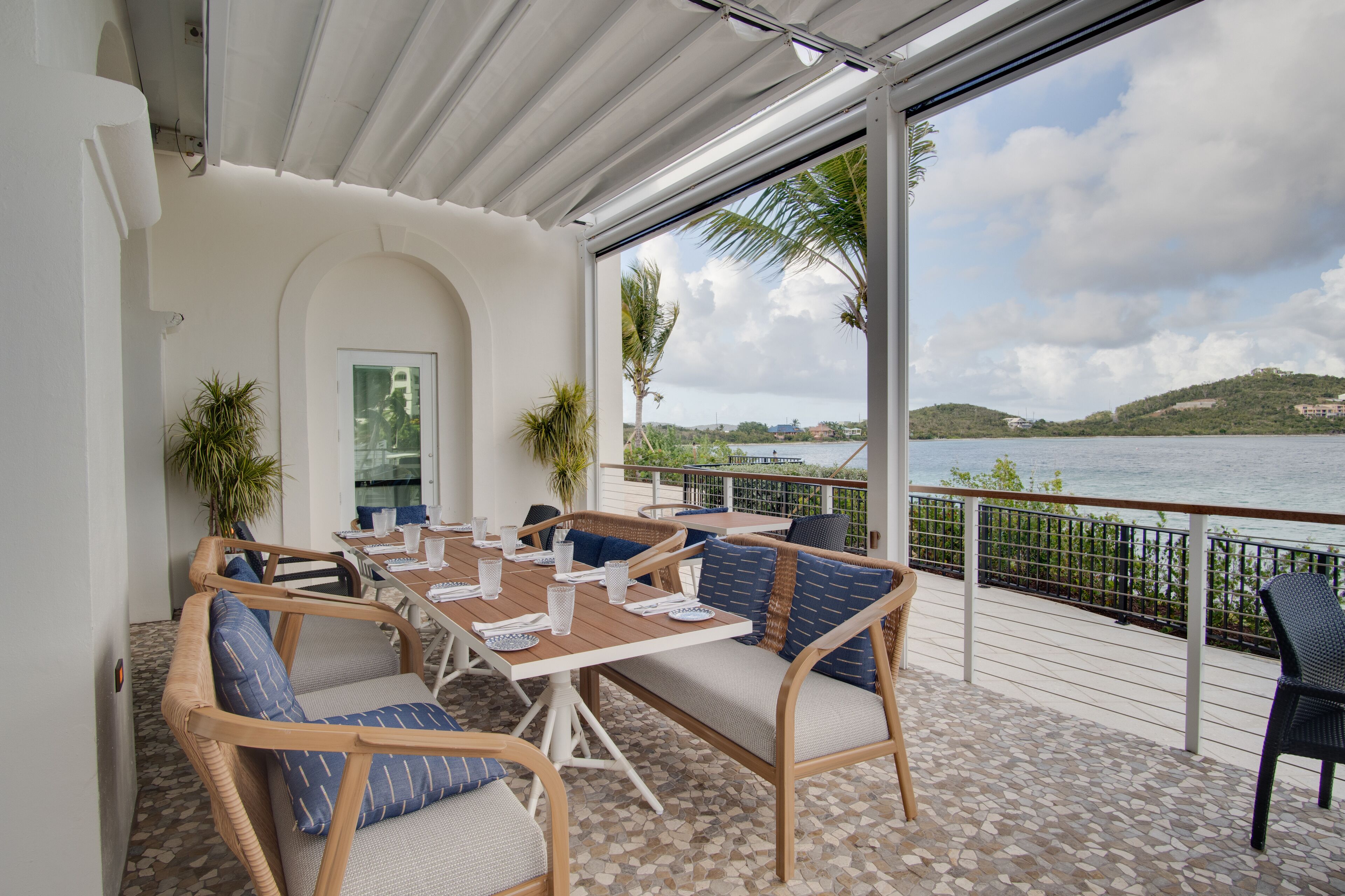 An outdoor dining area with wicker furniture, a tiled floor, and a view of the sea and hills.