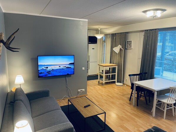 A modern living room with a grey sofa, a wooden coffee table, a TV mounted on the wall, a dining area with a white table and chairs, and a large window with curtains. - STAY Sello Business Suite (Espoo)