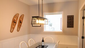A dining area with a wooden table and white chairs, a hanging light fixture, and a window with blinds. - Cozy ground level suite with private hot tub - close to the Attridge Chair (Silver Star Mountain)