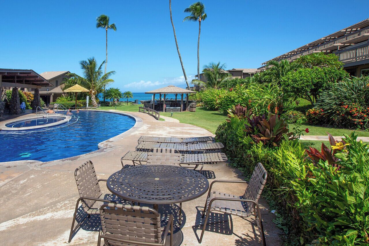 A pool area with lounge chairs and tables, surrounded by palm trees and a clear view of the ocean.
