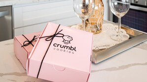 A kitchen with white cabinets, a stainless steel sink, and a microwave. There is a bottle of wine and two glasses on a tray. A pink box labeled "crumbl cookies" is also visible.