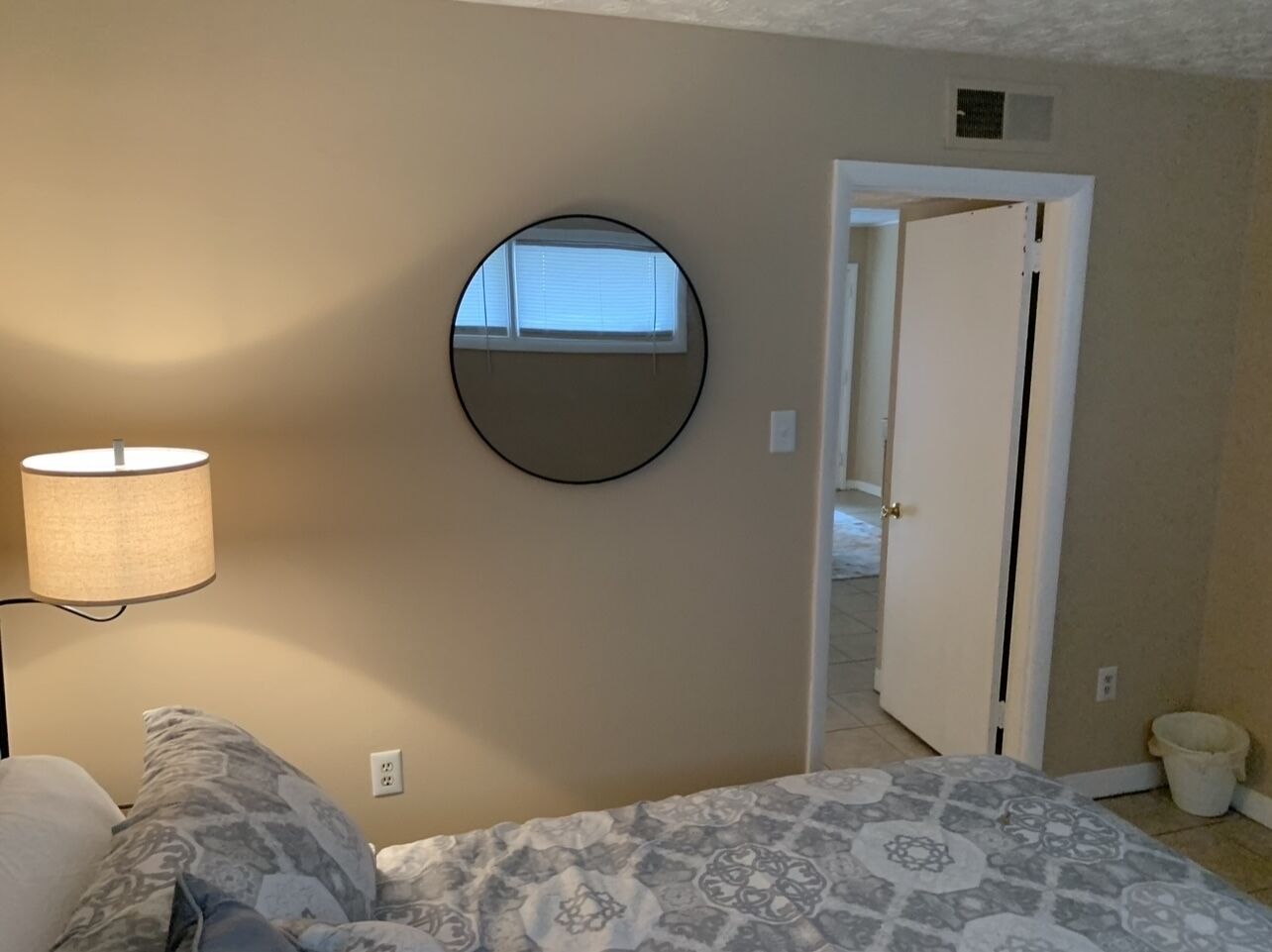 A bedroom with a bed, a round mirror, a lamp, and a door leading to another room.
