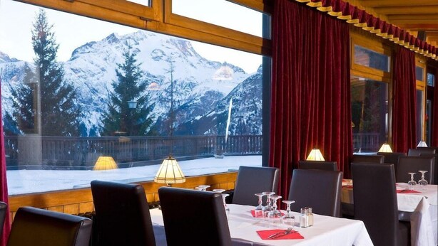 Ein Restaurant mit gedeckten Tischen, großen Fenstern mit Blick auf die Berge und roten Vorhängen.