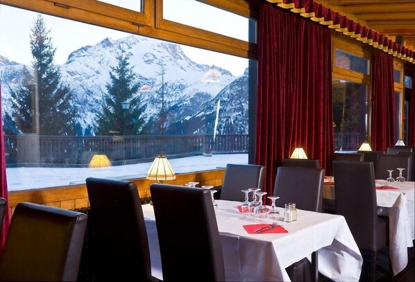 Ein Restaurant mit gedeckten Tischen, großen Fenstern mit Blick auf die Berge und roten Vorhängen.