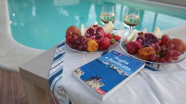 A poolside table with fruit, wine glasses, and a book titled 'Puglia e Basilicata'.