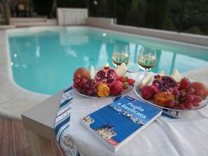 Una mesa junto a la piscina con fruta, copas de vino y un libro titulado 'Puglia e Basilicata'.