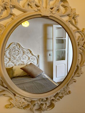 Features a bed with a decorative headboard and two pillows, reflected in a large ornate mirror.