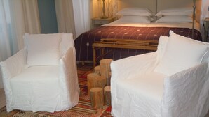A room with two white armchairs, a wooden bed with a patterned bedspread, and a colorful rug.