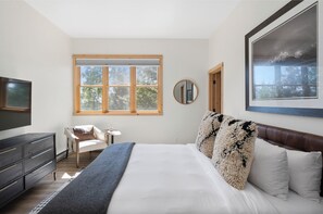A bedroom with a large bed, a TV, a chair, a round mirror, and a window with a view of trees. - Outpost: Moose Creek 34 - Private Hot Tub! (Teton Village)