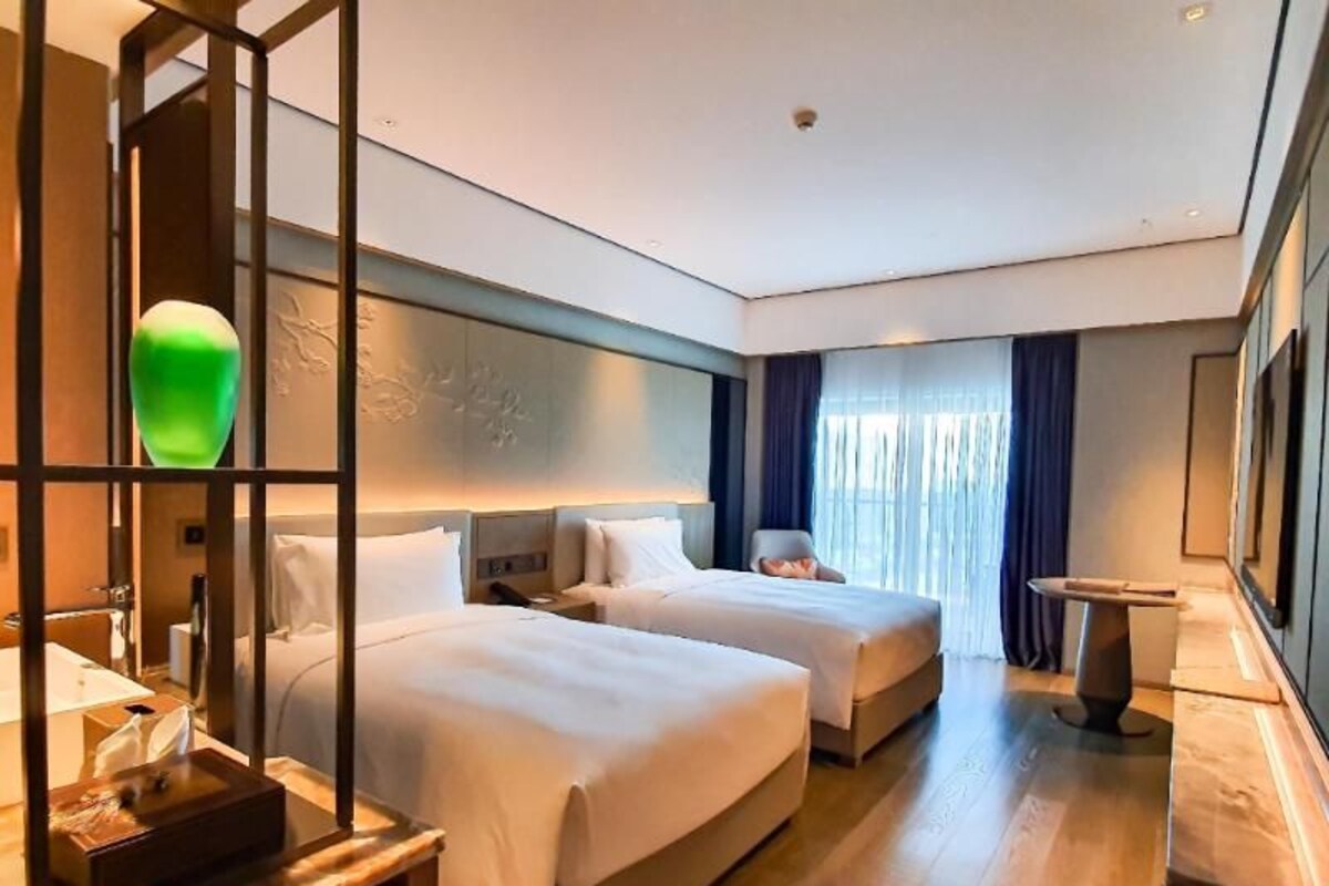 Habitación de hotel con dos camas, una mesita de noche con lámpara, una pequeña mesa y un amplio ventanal con cortinas.