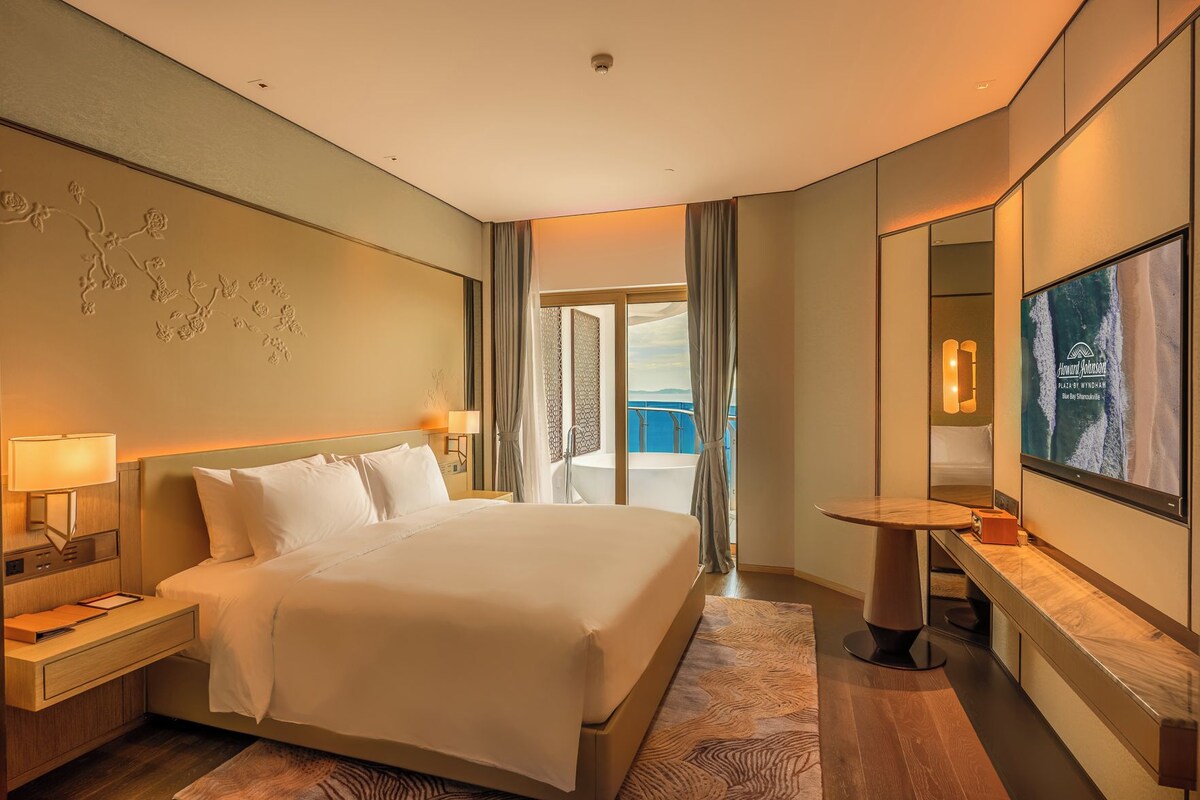 Habitación de hotel con una cama grande, un escritorio con lámpara, televisión y vistas al mar desde un balcón.