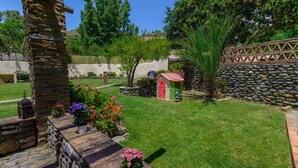 Un jardín bien cuidado con un muro de piedra, una casita de juegos roja y verde, y diversas plantas en flor.
