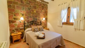 Un dormitorio con pared de piedra, una cama con colcha color beige, dos mesitas de noche con lámparas y una ventana con cortinas blancas.
