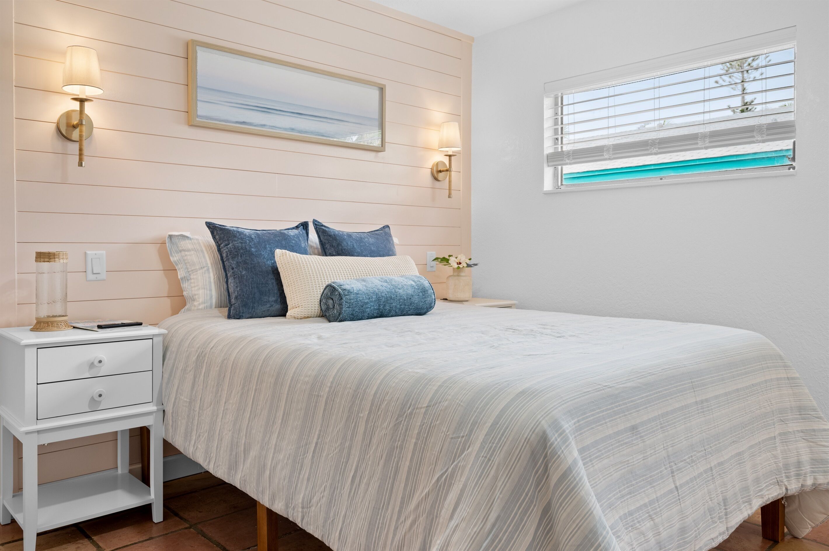 Une chambre à coucher comprenant un lit, une table de chevet, des lampes fixées au mur, une fenêtre avec des stores et un tableau encadré accroché au mur.