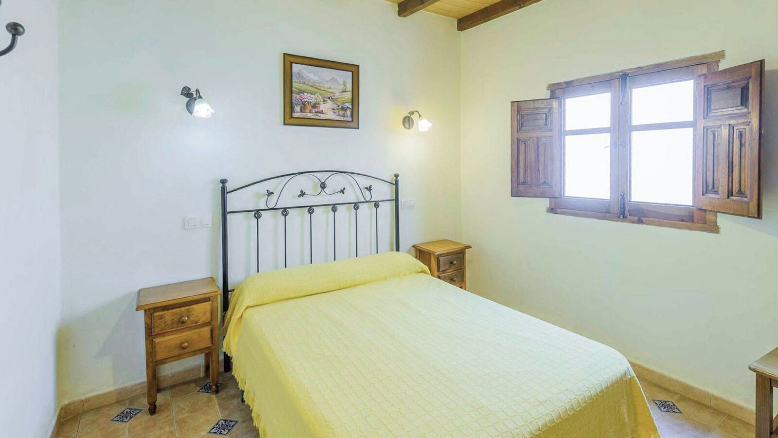 Ein Schlafzimmer mit einem Bett, zwei Nachttischen, einer Fensterbank, einem Fenster mit Holzläden und einer Wandleuchte.