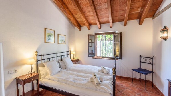 A bedroom with a wooden ceiling, a bed with white linens, a nightstand with a lamp, a chair, and a window with a view of greenery. - Finca Lomas de Tienda Antequera - La Higuera by Ruralidays (Málaga)
