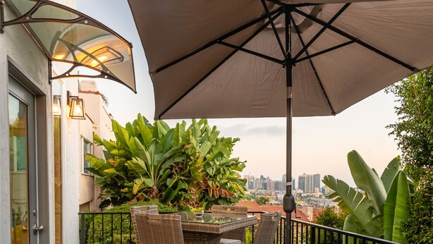 Eine Holzterrasse mit Tisch und Stühlen unter einem großen Sonnenschirm, umgeben von üppiger Bepflanzung und Blick auf die Stadt.