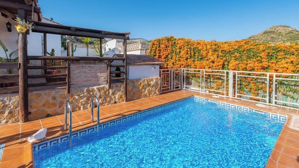 En swimmingpool med stenkant og fliser, omgivet af en træpergola og en hvid mur.