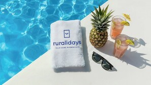 Una toalla con la marca 'ruralidays', una piña, dos bebidas con lima y gafas de sol junto a una piscina.