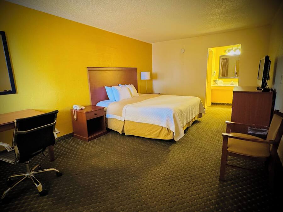Habitación de hotel con cama, escritorio, silla, mesita de noche y baño visible a través de una puerta abierta.