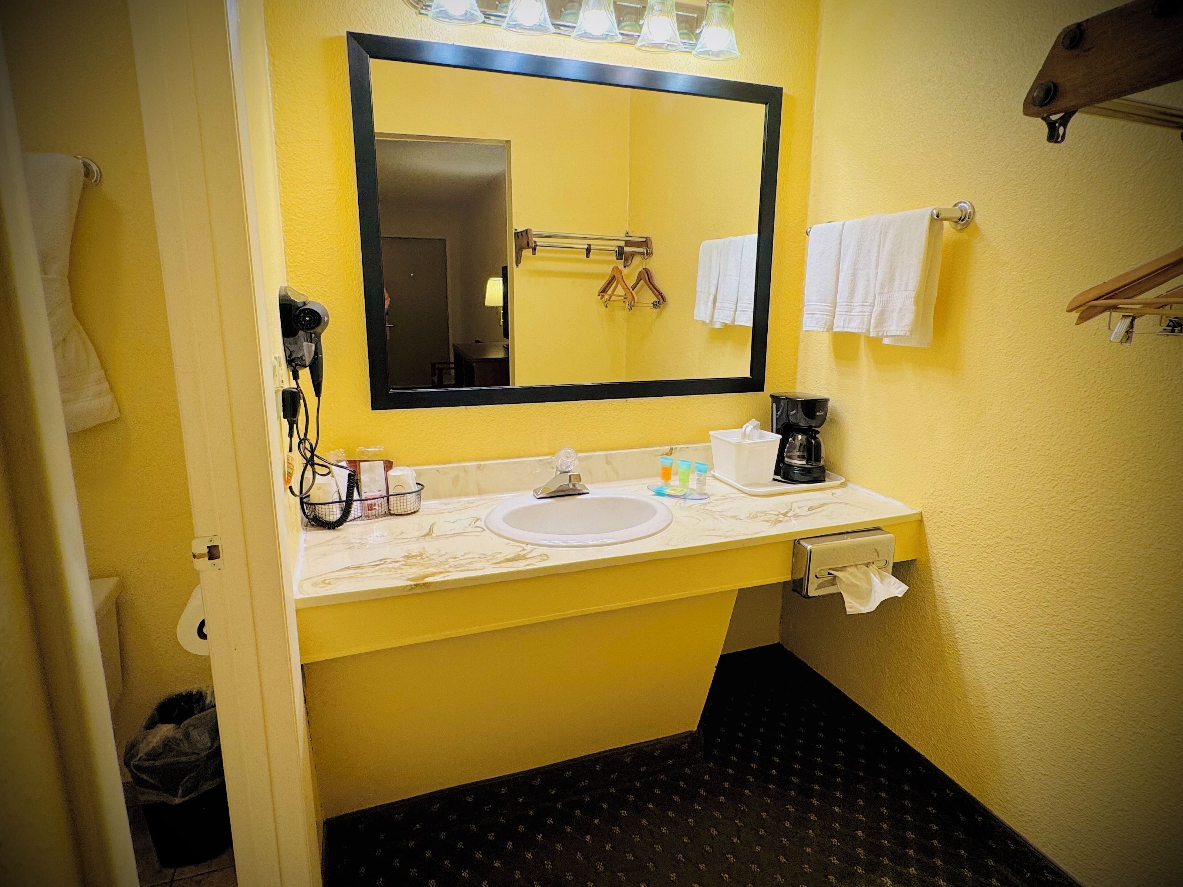 A bathroom with a yellow wall, a white sink, a mirror, a towel rack, and a coffee maker.
