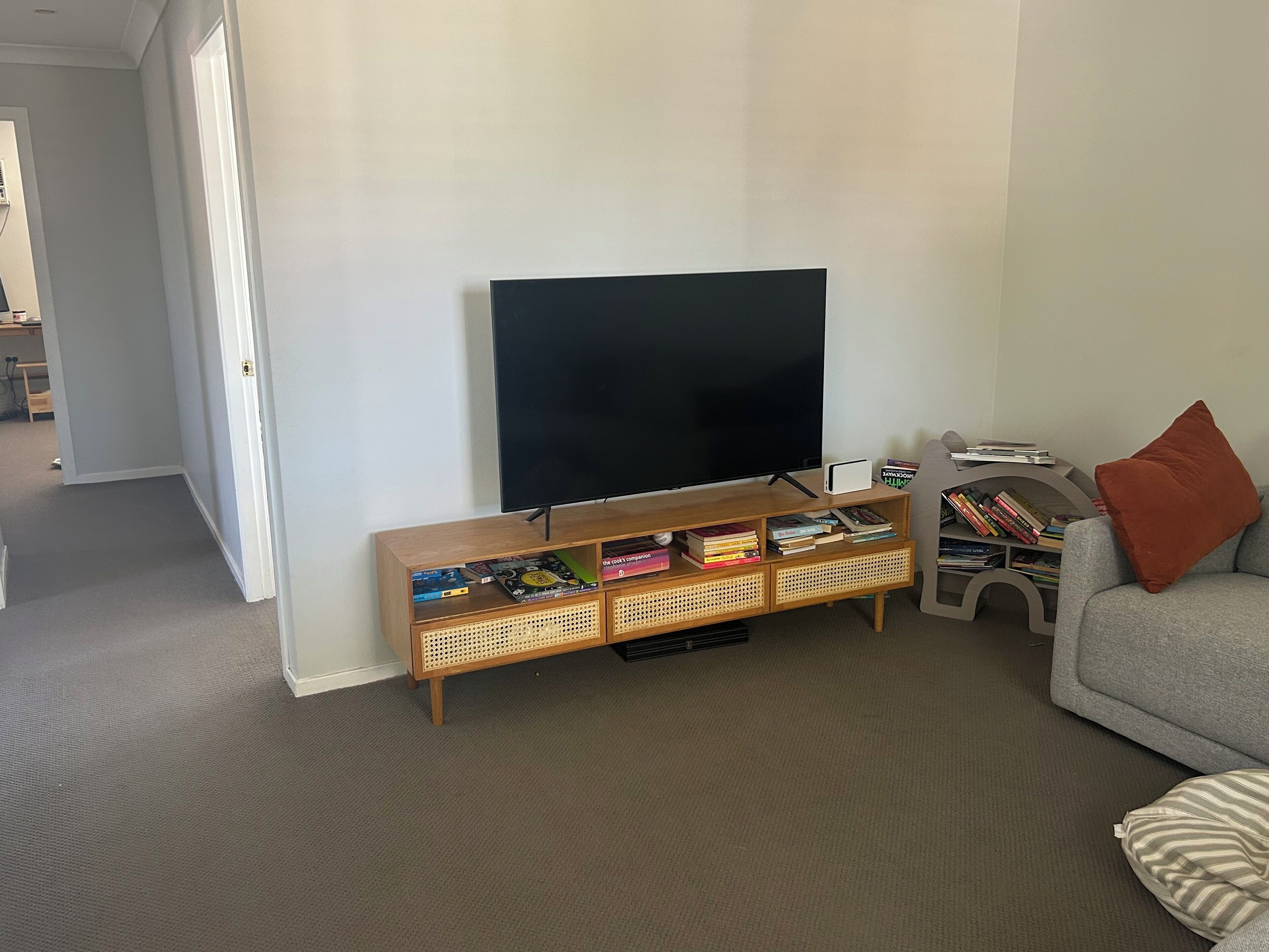 A living room with a flat-screen TV on a wooden stand, a grey sofa with a red pillow, and a bookshelf filled with books.