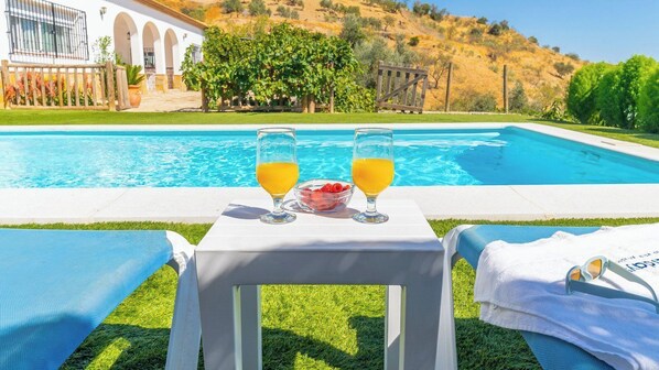 Une table au bord de la piscine avec deux verres de jus d’orange et un saladier de fraises, une serviette et des lunettes de soleil posées sur l’herbe.