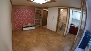A room with a wooden floor, a pink floral wall, a built-in shelf unit, a door leading to a bathroom, and a kitchen area visible through an open door.