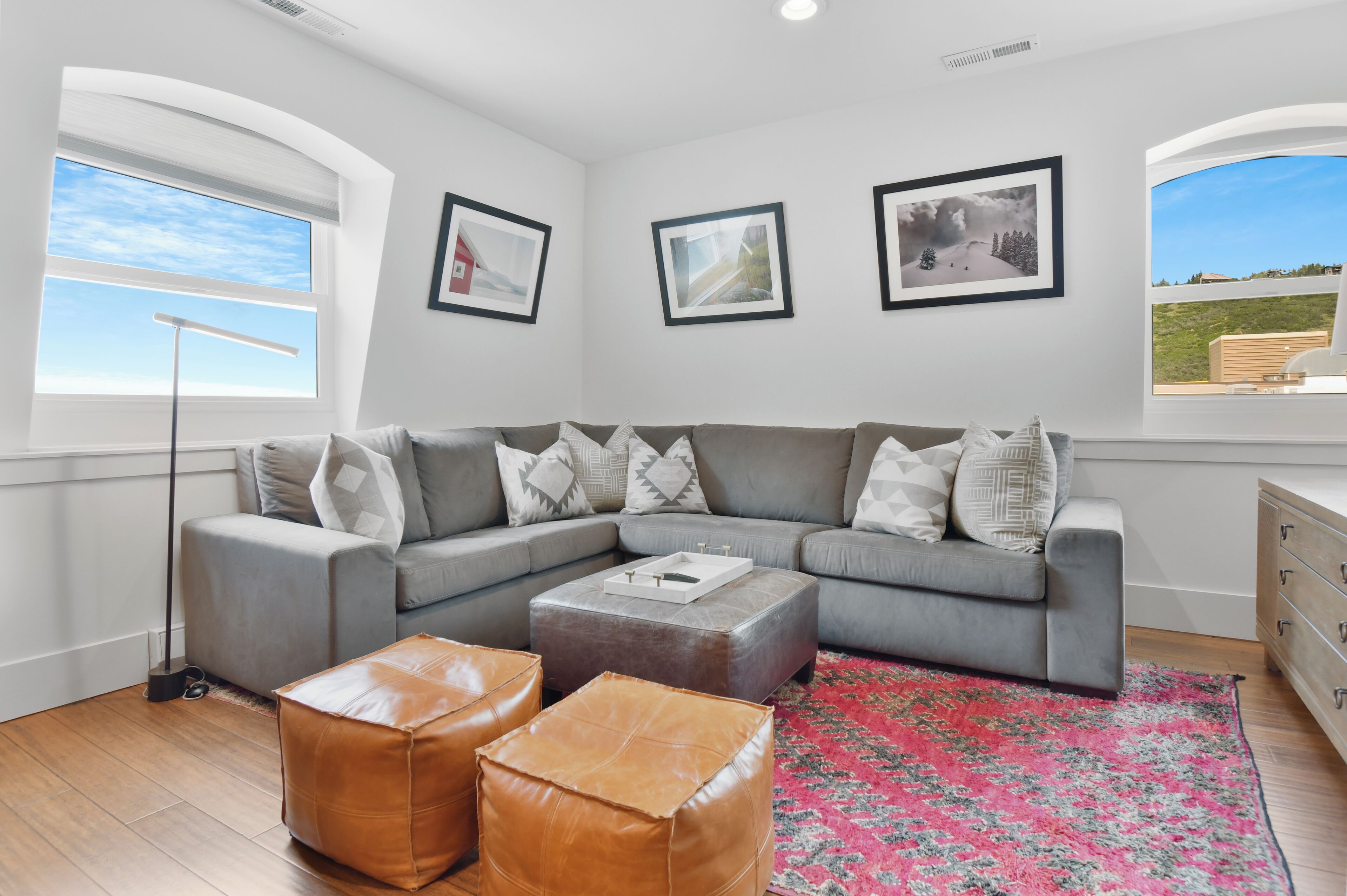 A modern living room with a grey sectional sofa, a small ottoman, a floor lamp, and a red and grey patterned rug. There are three framed pictures on the wall and a view of a landscape through the window.