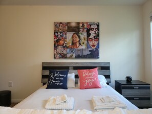 Una camera d'albergo con un letto, due cuscini con scritte, un'opera d'arte a parete e un comodino con una sveglia.