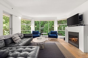 A modern living room with a fireplace, leather sofa, blue armchairs, and large windows offering a view of greenery. - Mountain condo w/ pool & hot tub (Whistler)
