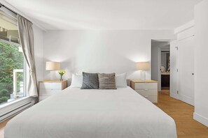 A bedroom with a large bed, two bedside tables with lamps, a window with curtains, and a view of greenery outside. - Mountain condo w/ pool & hot tub (Whistler)