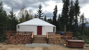 A single-story yurt with a red door, wooden steps leading up to it, and stacks of firewood on either side.
