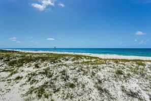 Une plage de sable blanc avec une végétation clairsemée, un ciel bleu azur et un voilier à l’horizon.