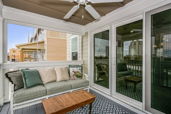 A covered balcony with a sofa, a wooden table, and a view of a cityscape.
