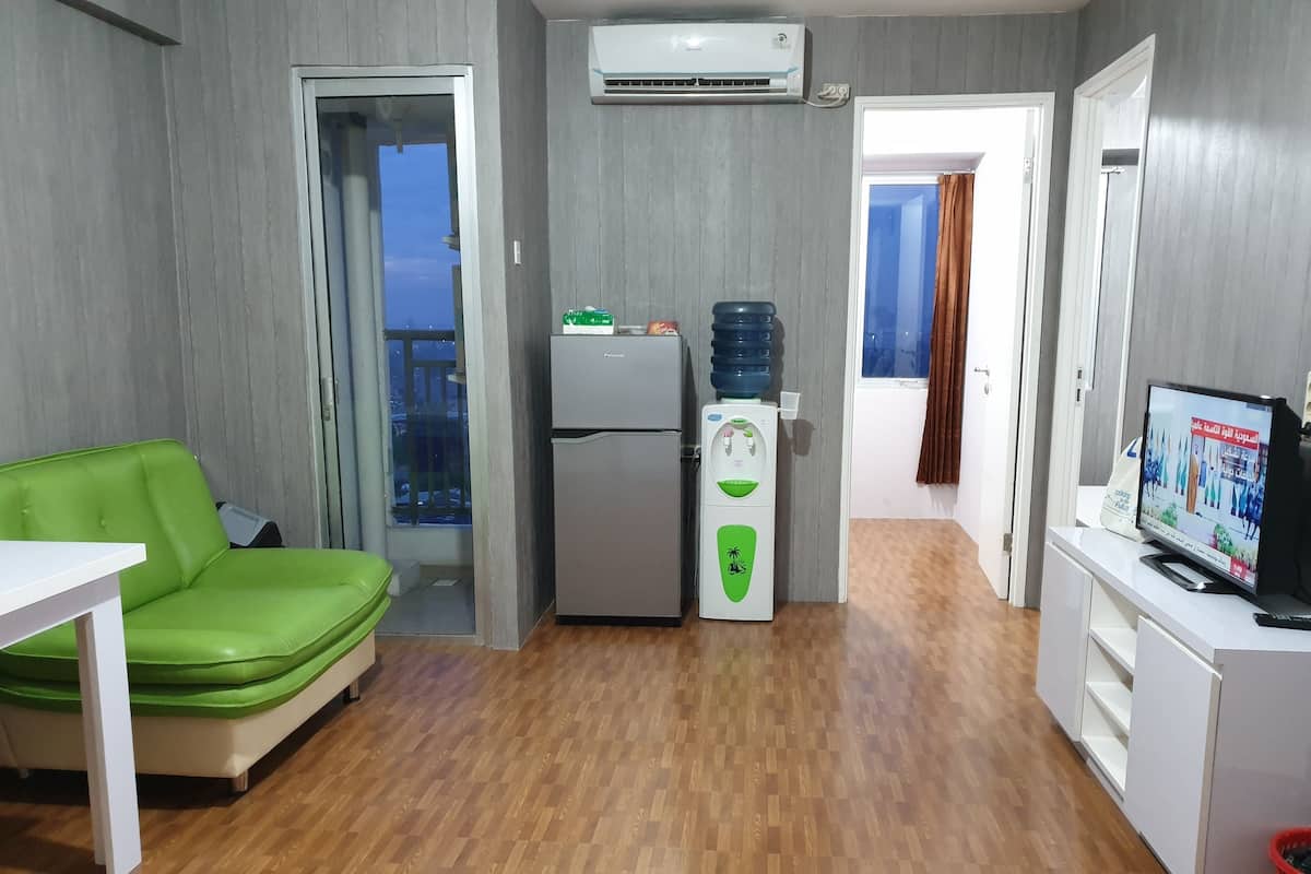 Un espace de vie compact comprenant un canapé vert, un mini-réfrigérateur, un distributeur d’eau, une télévision et un balcon avec vue.