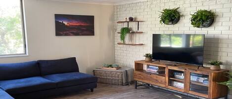 A living room with a dark blue sofa, a flat-screen TV on a wooden stand, a white brick wall, and two potted plants.