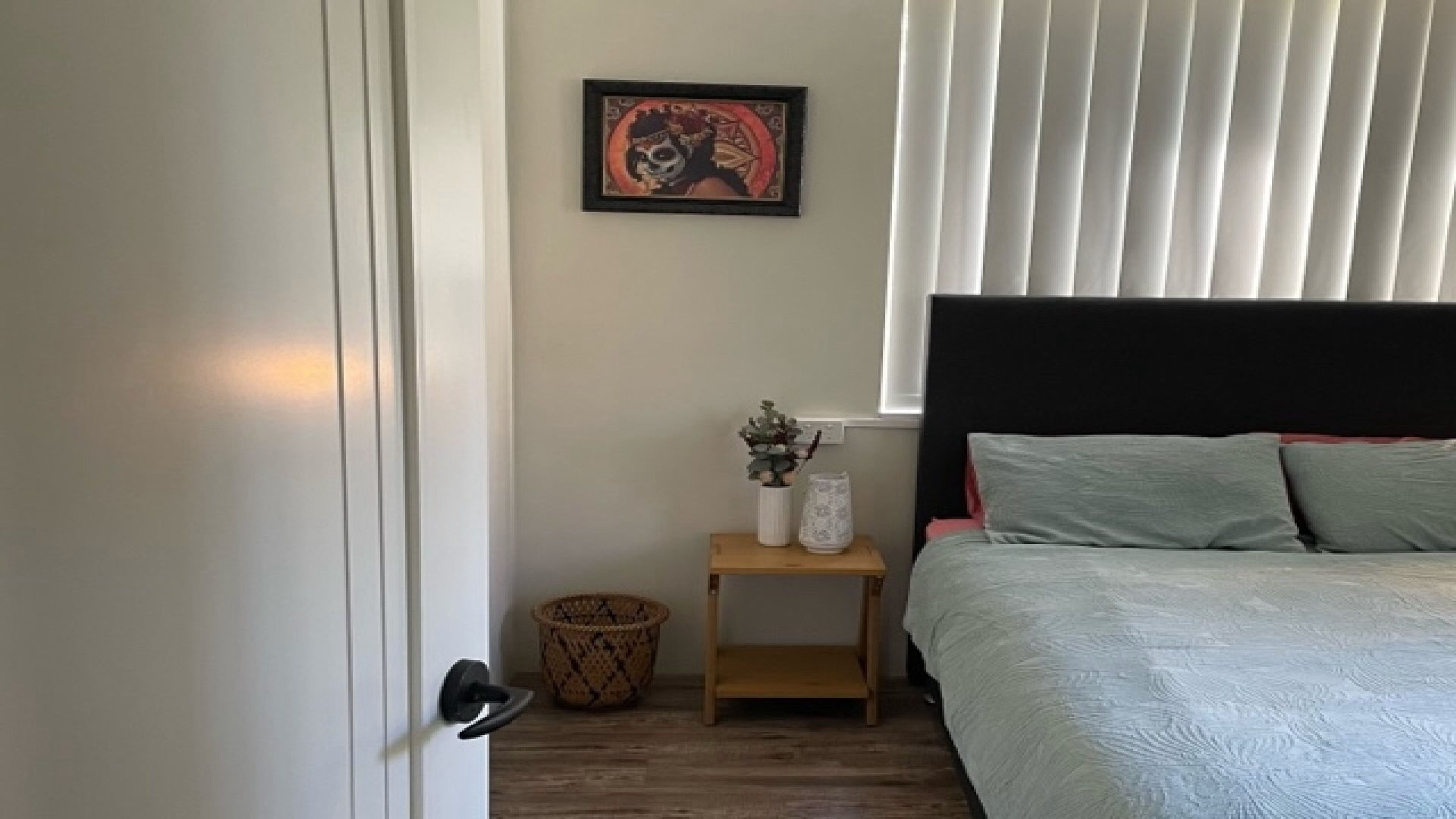 Quarto com cama, mesa de cabeceira, quadro emoldurado e janela com persianas.