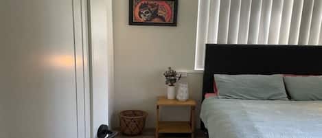 Quarto com cama, mesa de cabeceira, quadro emoldurado e janela com persianas.