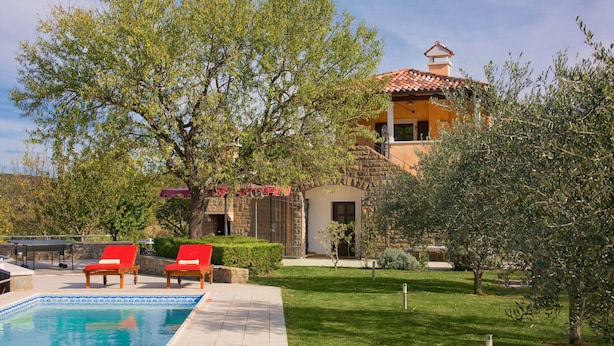 A pool area with two red lounge chairs, a stone wall, and a building with a tiled roof in the background.
