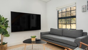Ett modernt vardagsrum med en stor platt-TV som är monterad på väggen, en grå soffa, ett litet runt soffbord med en växt och ett fönster med utsikt över träd.