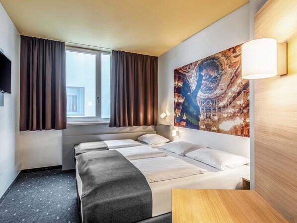 Una habitación de hotel con dos camas, un ventanal grande con cortinas, un televisor montado en la pared y un tapiz de gran tamaño.