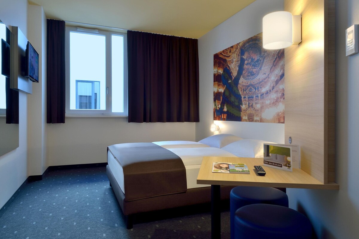 A hotel room with a bed, a desk with a remote control, a lamp, and a painting on the wall.