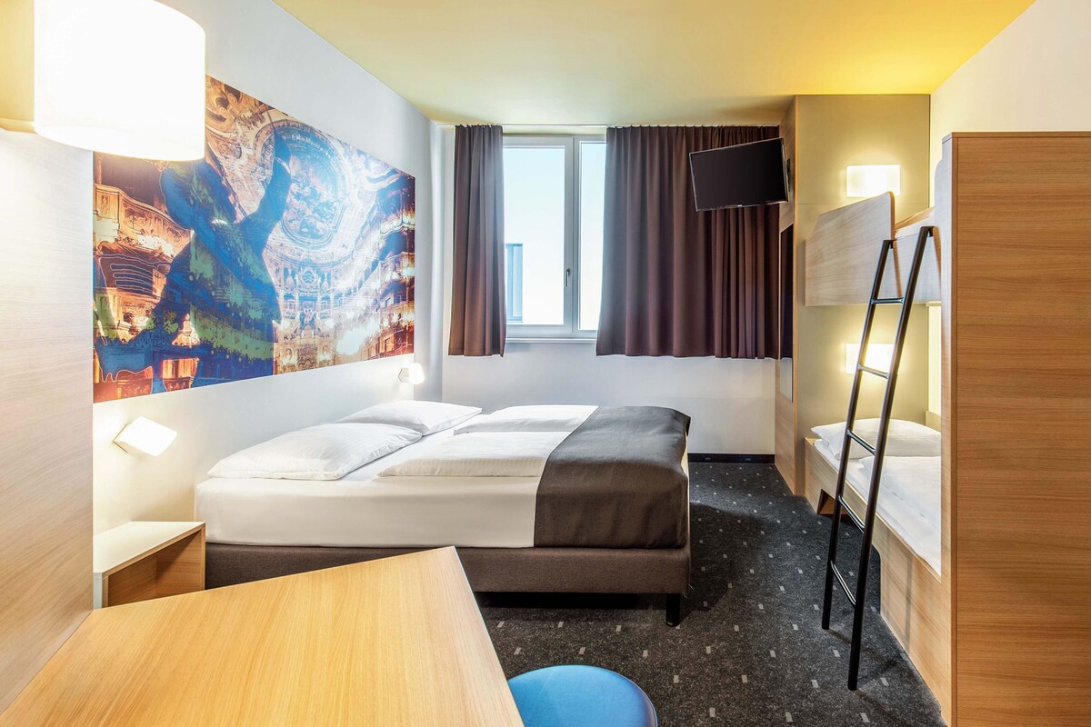 Ein Hotelzimmer mit Bett, Schreibtisch, Stuhl, Fernseher und einem Fenster mit Vorhängen.