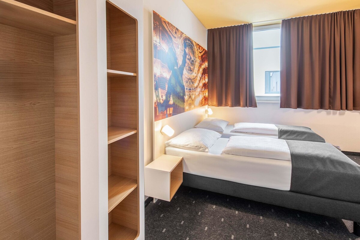 Ein Hotelzimmer mit einem Bett, einem Kopfende mit Kunstwerk, einem Kleiderschrank und einem Fenster mit Vorhängen.