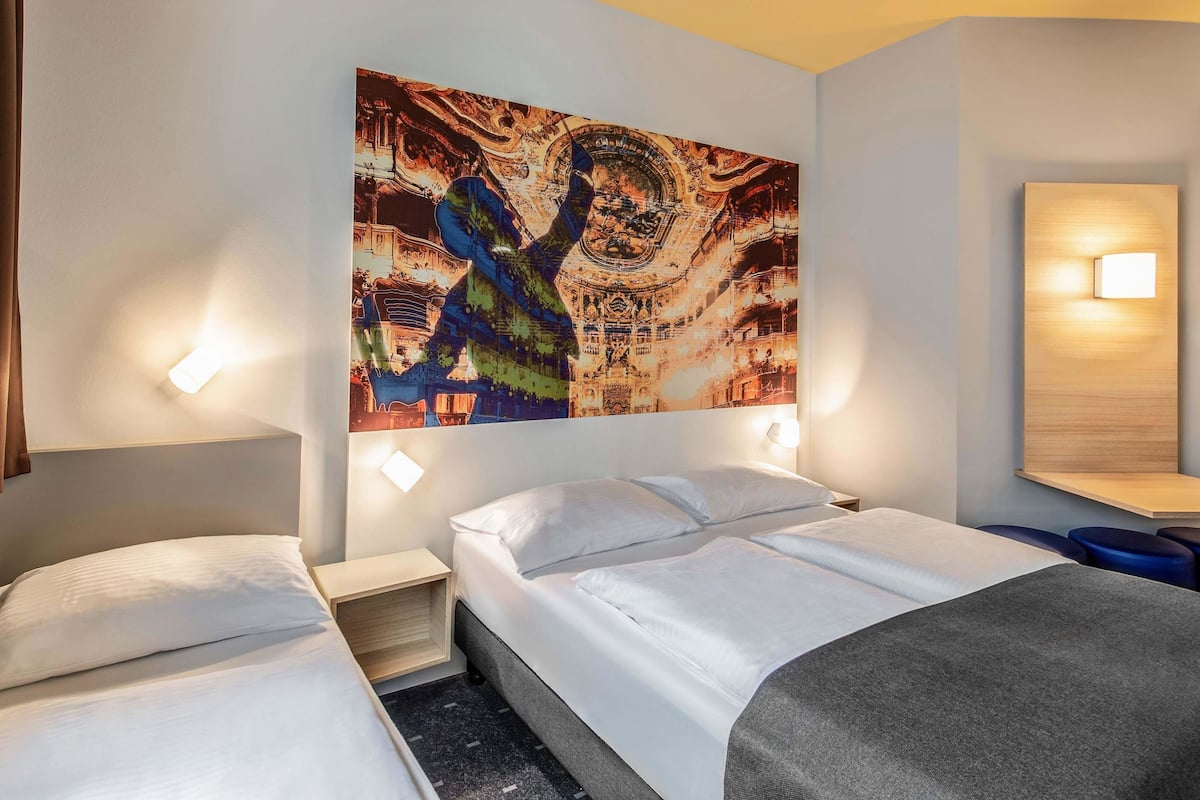 A hotel room with two beds, a large abstract painting on the wall, and a nightstand with a lamp.